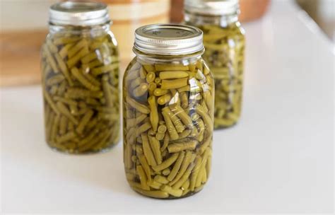 The process of canning green beans, showing washed beans in a jar and other ingredients on a kitchen counter