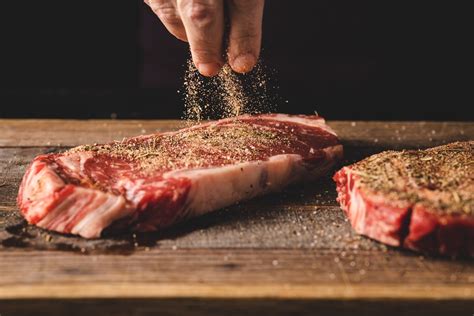 Close-up photo of raw meat being seasoned with salt, pepper, rosemary, garlic, and onion slices.
