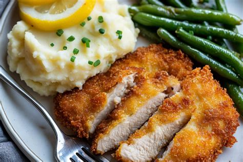 Thematic photo of a plate with a crispy pork schnitzel, oven-baked potatoes, and a fresh green salad. Garnished with a lemon wedge and a sprig of rosemary.