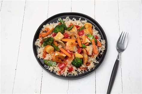 A beautifully plated dish of sweet and sour chicken with various colorful vegetables and rice, garnished with sesame seeds and coriander.